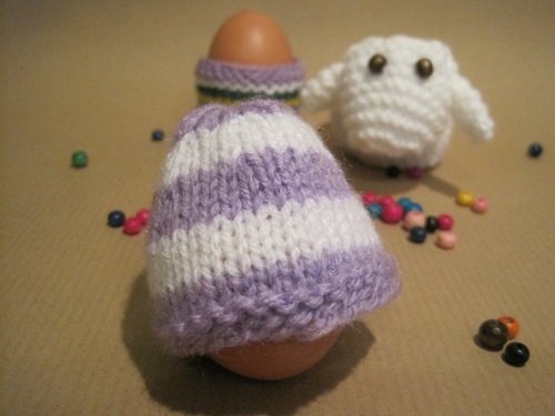 Zwei Eier mit gestrickten Eierwärmern (lila-weiß gestreift), kleine weiße Strickfigur und bunte Perlen
