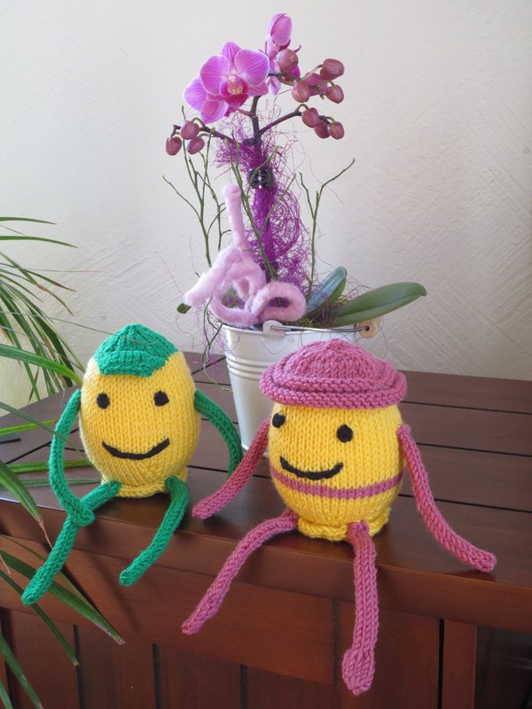 Zwei gelbe gestrickte Ostereier mit lächelnden Gesichtern auf einem Holzsideboard vor einer violetten Orchidee