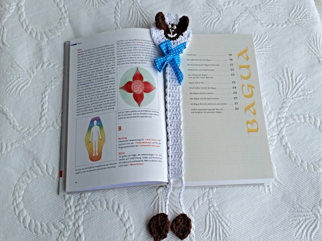 Weißes gehäkeltes Lesezeichen mit braun-weißem Schafkopf, blauer gepunkteter Schleife und zwei gehäkelten Endstücken in einem geöffneten Buch