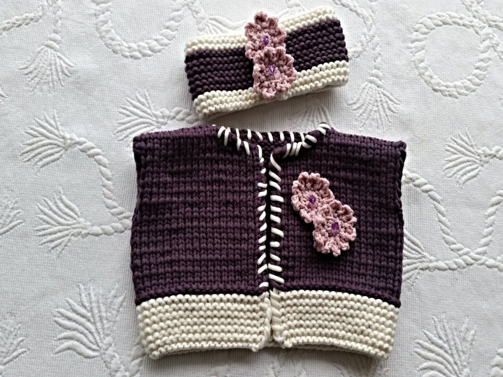 Gestrickte ärmellose Babyweste und Stirnband in Lila und Creme mit rosa Blumenapplikationen.