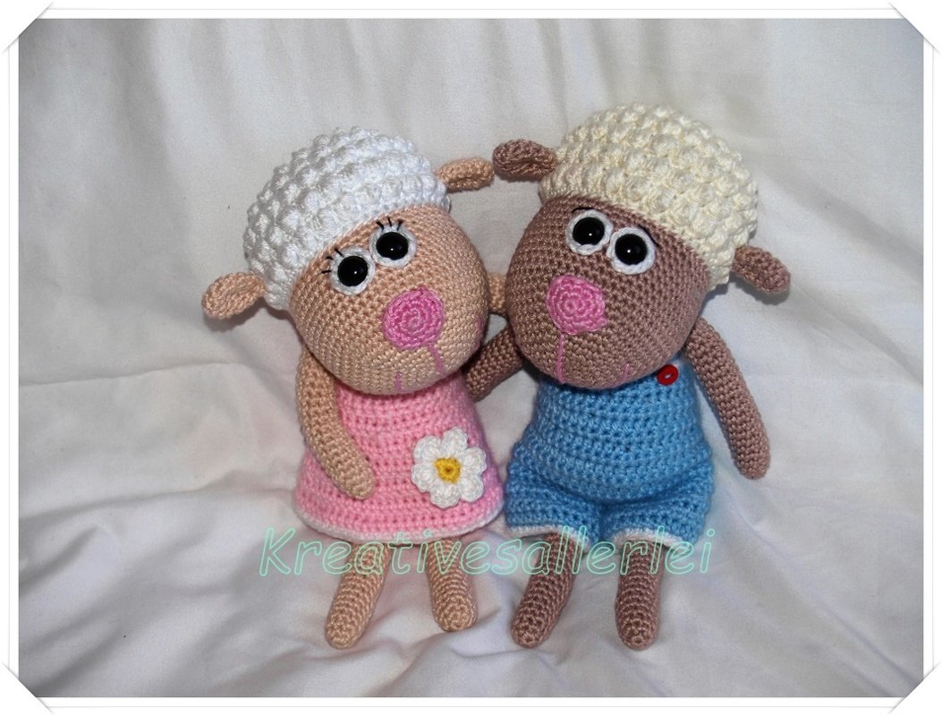 Zwei gehäkelte Amigurumi-Schafe, eins in rosa Kleid mit weißer Blume, eins in blauer Latzhose, beide mit strukturierter Kopfform und großen Augen