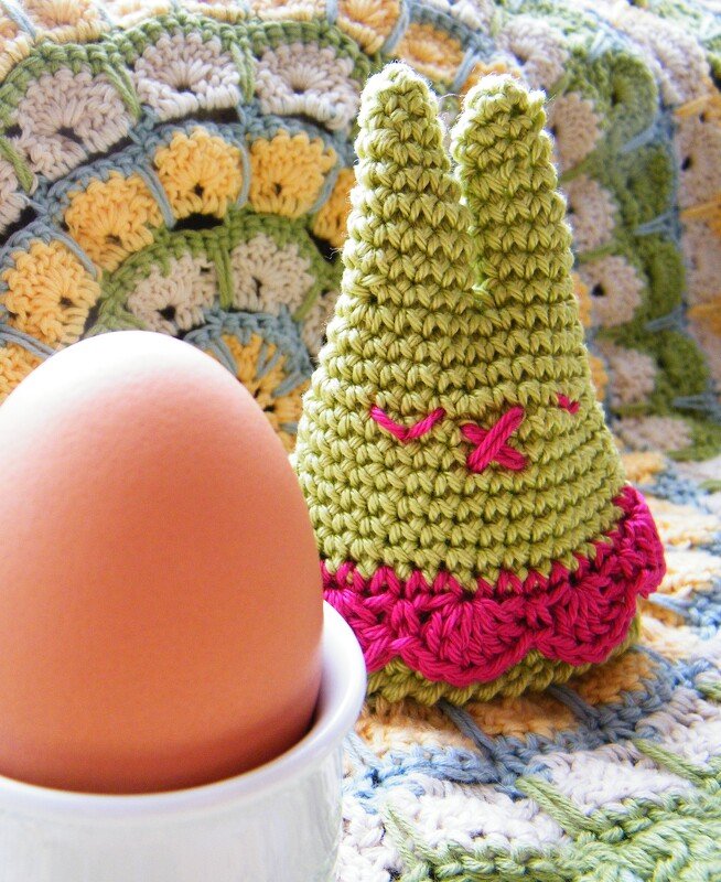 Gehäkelter grüner Hasen-Eierwärmer mit rosa Verzierungen neben einem Ei im weißen Eierbecher auf bunter Häkeldecke.
