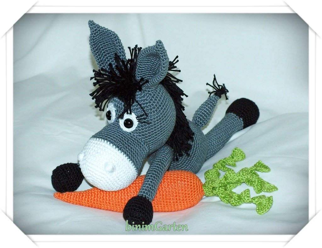 Gehäkelter grauer Amigurumi-Esel liegt auf einer orangefarbenen gehäkelten Karotte