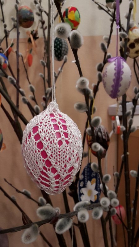 Pinkes Osterei in weiß gestrickter Spitzenhülle an Weidenkätzchenzweig, im Hintergrund weitere dekorierte Eier