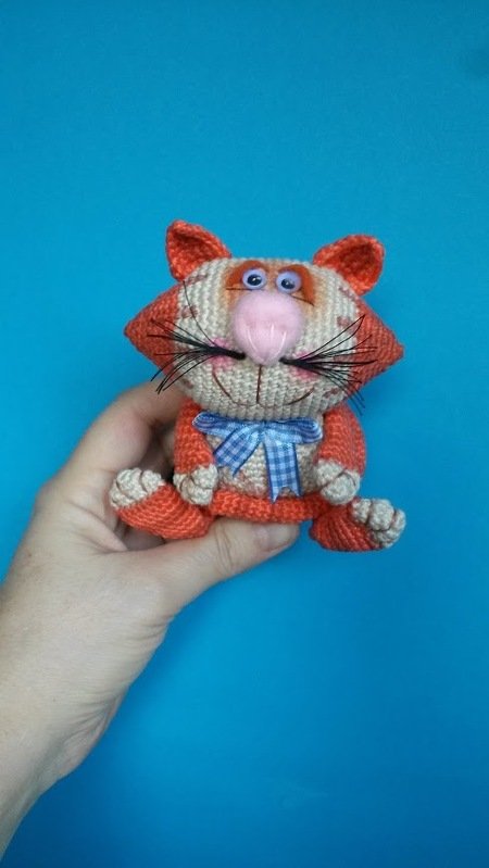 Hand hält gehäkelte Amigurumi-Katze in Orange und Creme mit rosa Nase und blau kariertem Schleifchen vor türkisfarbenem Hintergrund