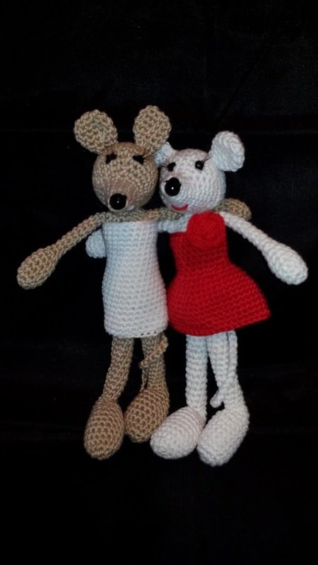 Zwei gehäkelte Amigurumi-Mäuse, eine beige mit weißem Kleid und eine weiße mit rotem Kleid, Arm in Arm vor schwarzem Hintergrund