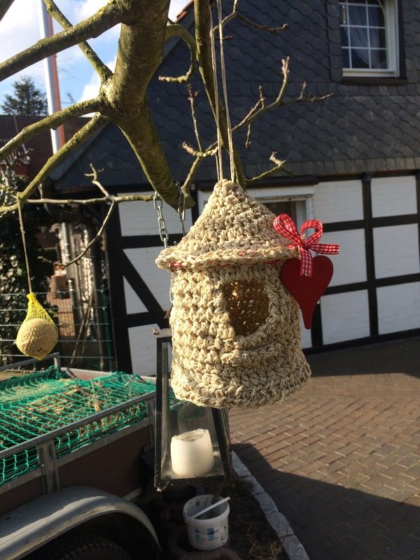 Beige gehäkeltes Vogelhäuschen mit rundem Loch und rotem Herz an einem Ast vor einem Fachwerkhaus