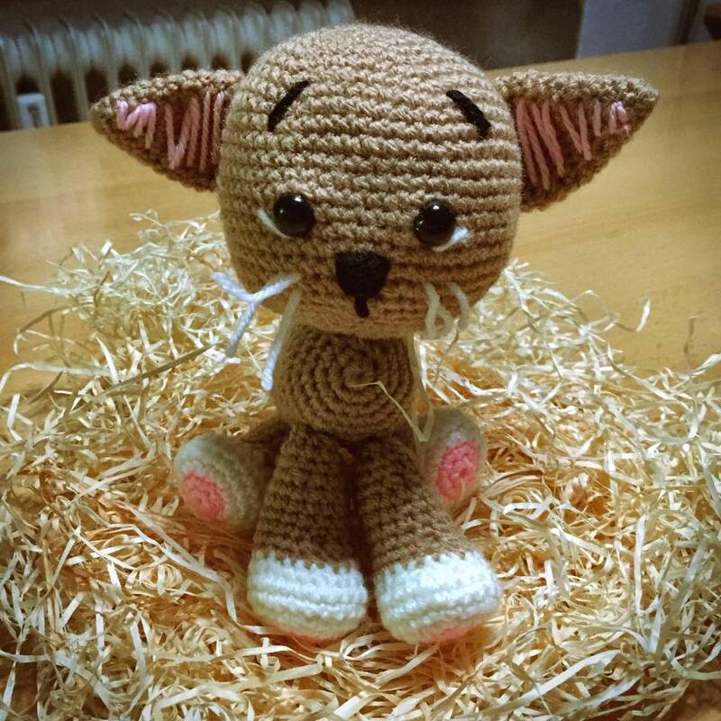 Braune gehäkelte Amigurumi-Katze mit weißen Pfoten und rosa Innenohren, sitzt auf Holzwolle