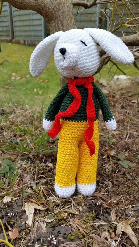 Weißer gehäkelter Amigurumi-Hase mit grünem Pullover, rotem Schal und gelber Hose vor einem Baumstamm