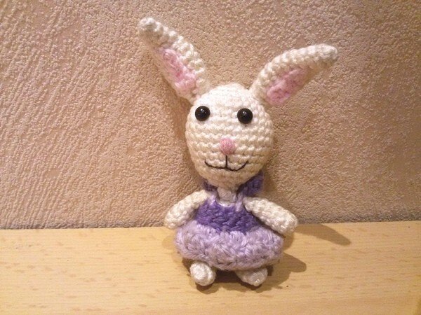 Weißer gehäkelter Amigurumi-Hase mit lila Kleid, rosa Innenohren und schwarzen Knopfaugen
