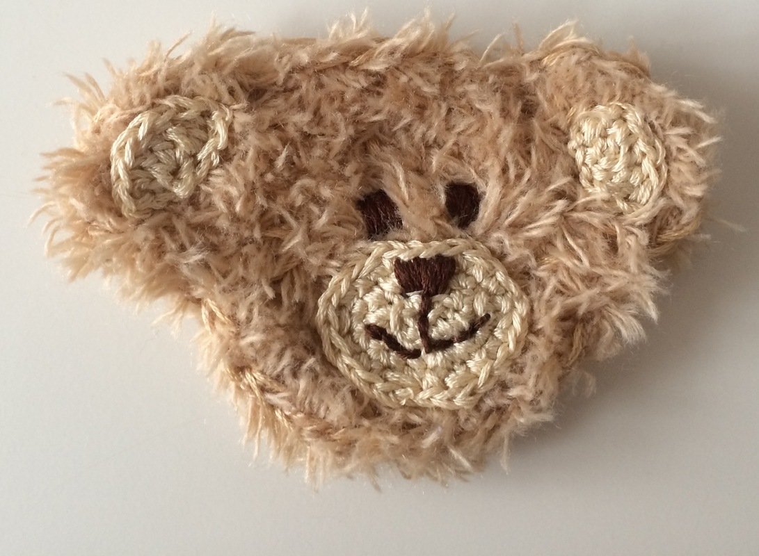 Flauschige, beige gehäkelte Teddykopf-Applikation mit gesticktem Gesicht.