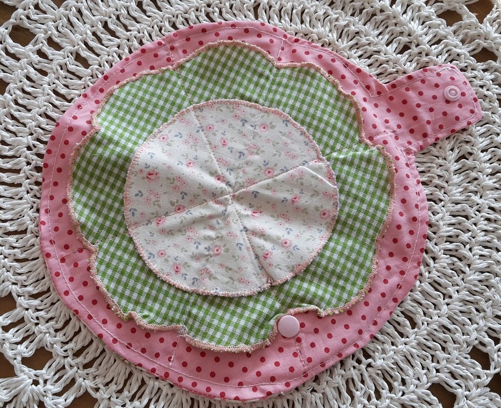 Rosa gepunktetes Stoff-Utensilo in Blütenform mit grünem Vichy-Karo und Blumenmuster auf weißer Häkeldecke
