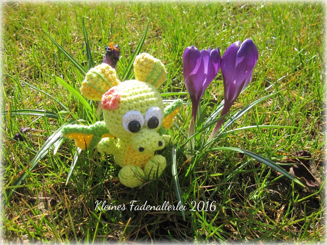 Gehäkelter hellgrüner und gelber Amigurumi-Drache mit großen Augen im Gras neben violetten Krokussen.