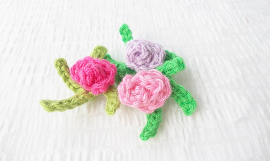 Drei gehäkelte Rosen-Applikationen in Pink, Hellrosa und Flieder mit grünen Blättern auf weißem Hintergrund