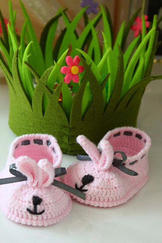 Rosa gehäkelte Babyschuhe mit Hasenohren und grauen Schleifen vor grüner Filzgras-Dekoration mit Blumen