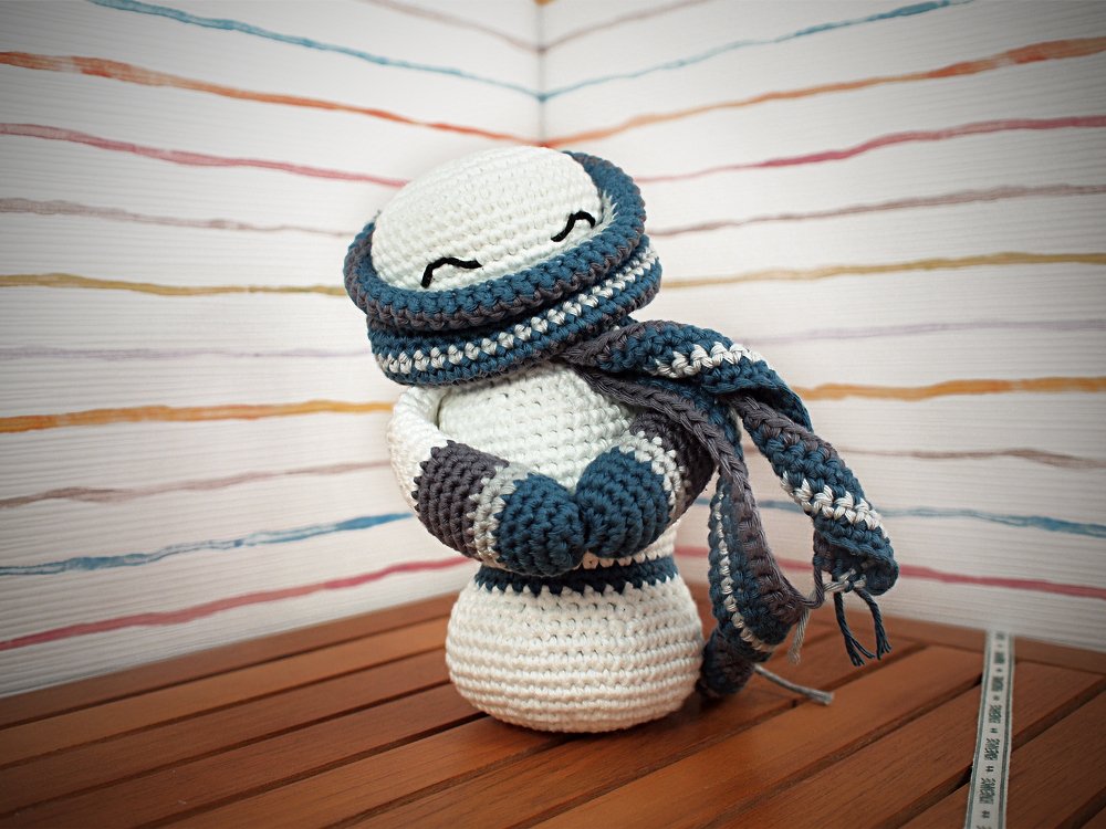 Gehäkelter weißer Amigurumi‑Schneemann mit blau‑grau‑weiß gestreiftem Schal auf Holzfläche