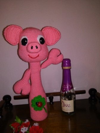 Rosa gehäkelter Flaschenüberzug in Form eines Glücksschweins mit Kleeblatt und Marienkäfer neben einer Flasche.