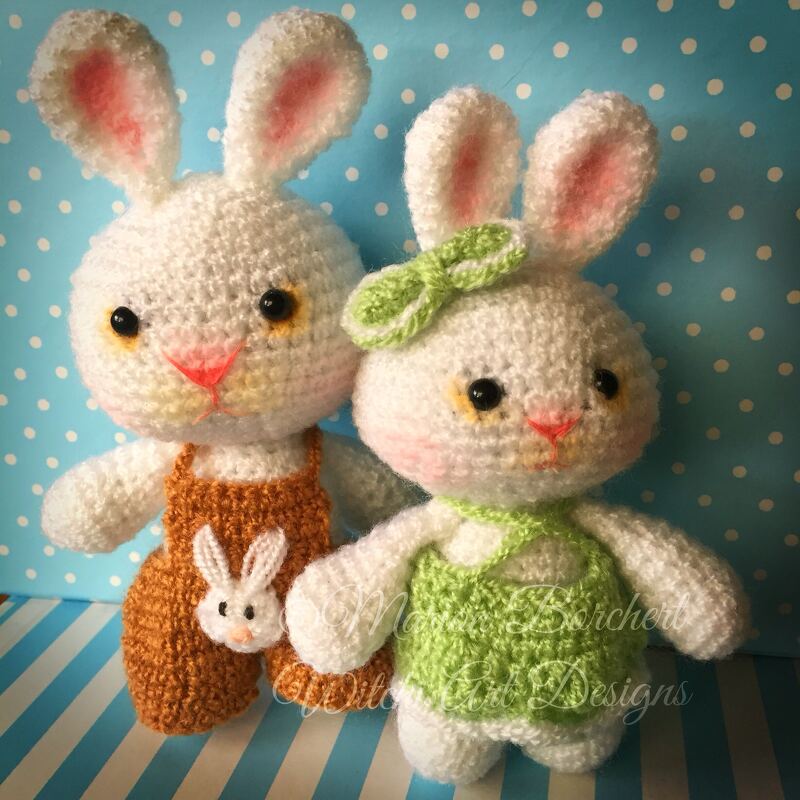 Zwei weiße gehäkelte Amigurumi-Hasen, einer mit brauner Latzhose und Hasenapplikation, einer mit grünem Kleid und Schleife vor hellblauem Punkt-Hintergrund