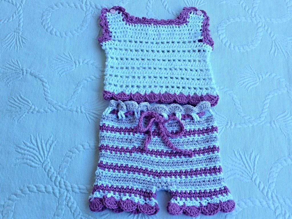 Zweiteiliges gehäkeltes Baby-Set in Weiß und Lila: ärmelloses Oberteil mit Knöpfen an den Schultern und gestreifte Shorts mit Kordelzug, auf hellblauem Untergrund.