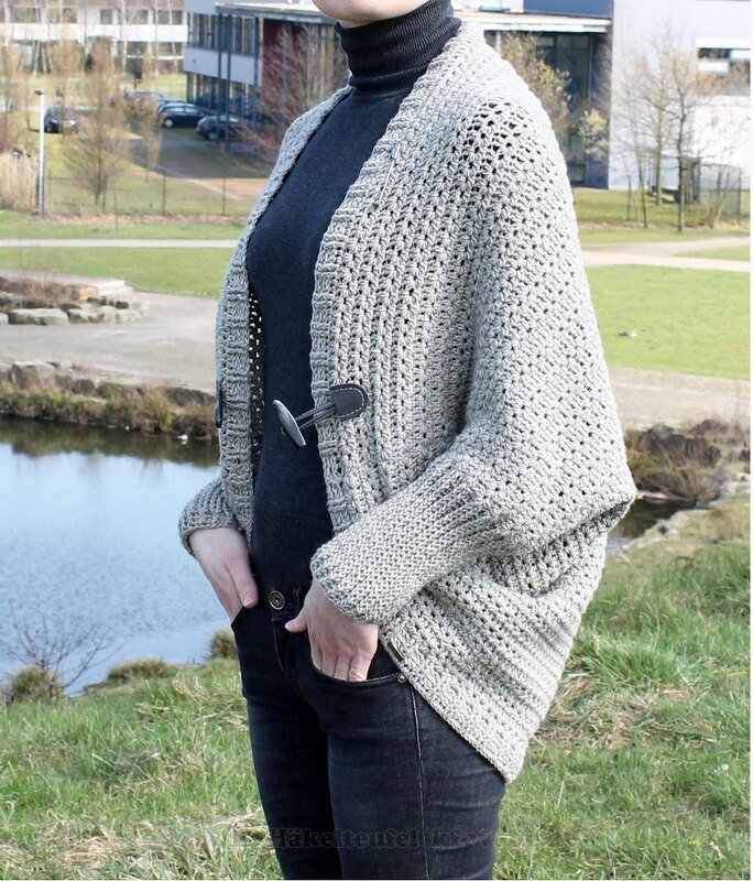 Person trägt hellgraue gehäkelte Oversize-Jacke mit Knebelverschluss vor einem See.