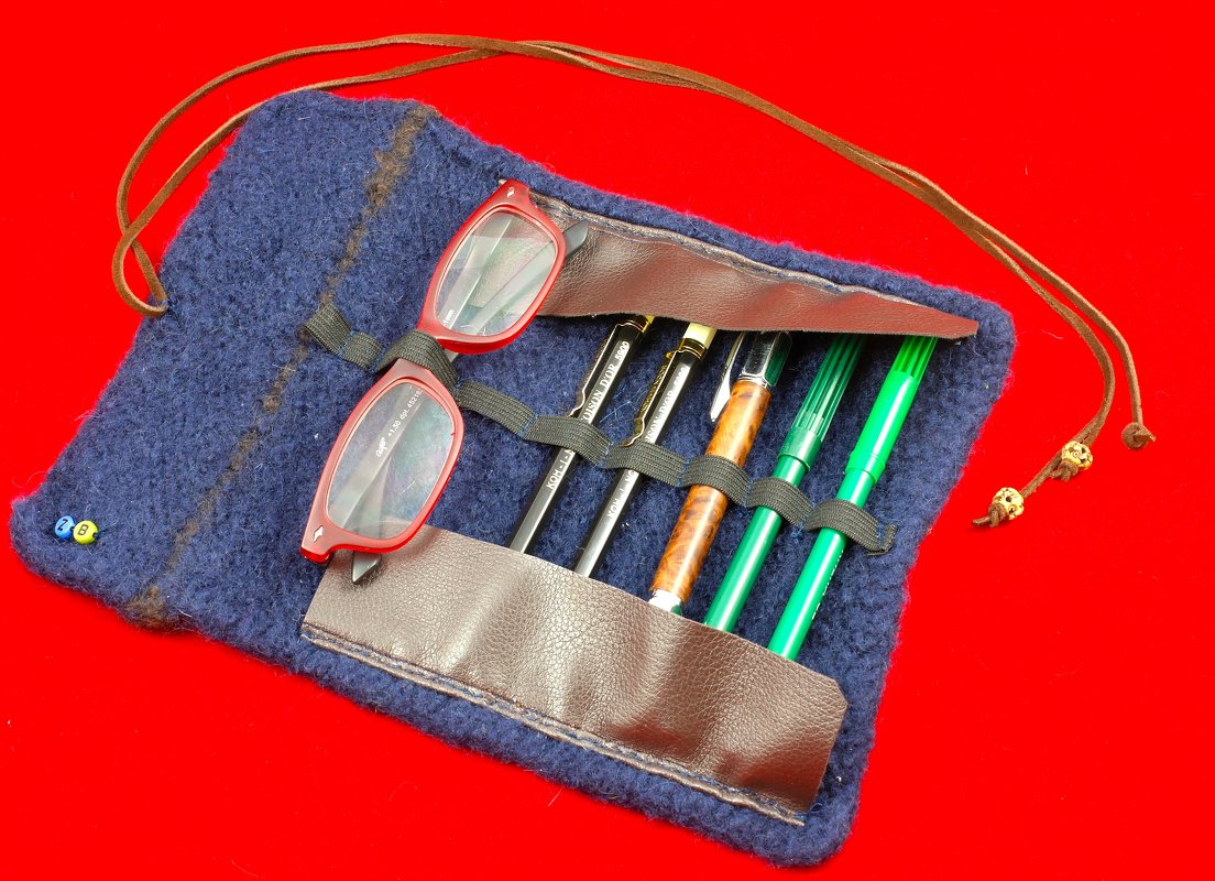 Blaues gestricktes, gefilztes Rollenetui mit brauner Lederklappe, elastischen Schlaufen, mehreren Stiften und roter Brille auf rotem Hintergrund