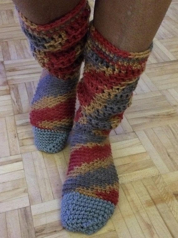 Close-up of legs wearing colorful striped crocheted socks with scrunched legwarmer-style cuffs on a wooden floor.