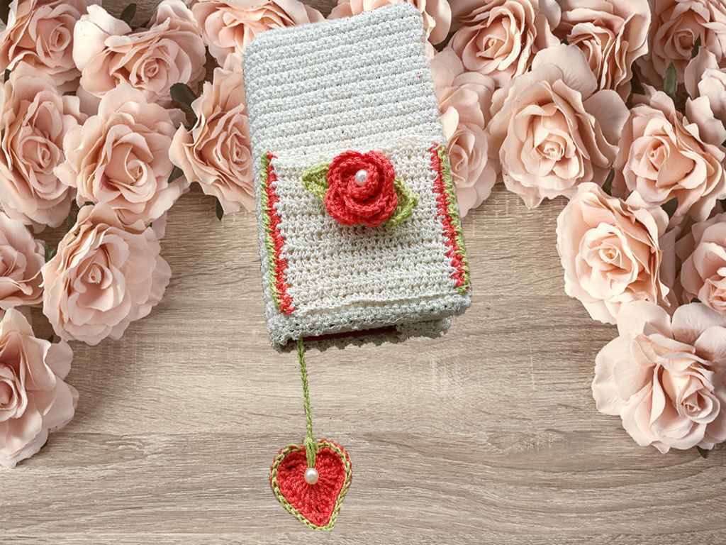 Cremefarbene gehäkelte Hülle mit roter Rose und Herzanhänger auf einem Holztisch, umgeben von rosafarbenen Rosen.