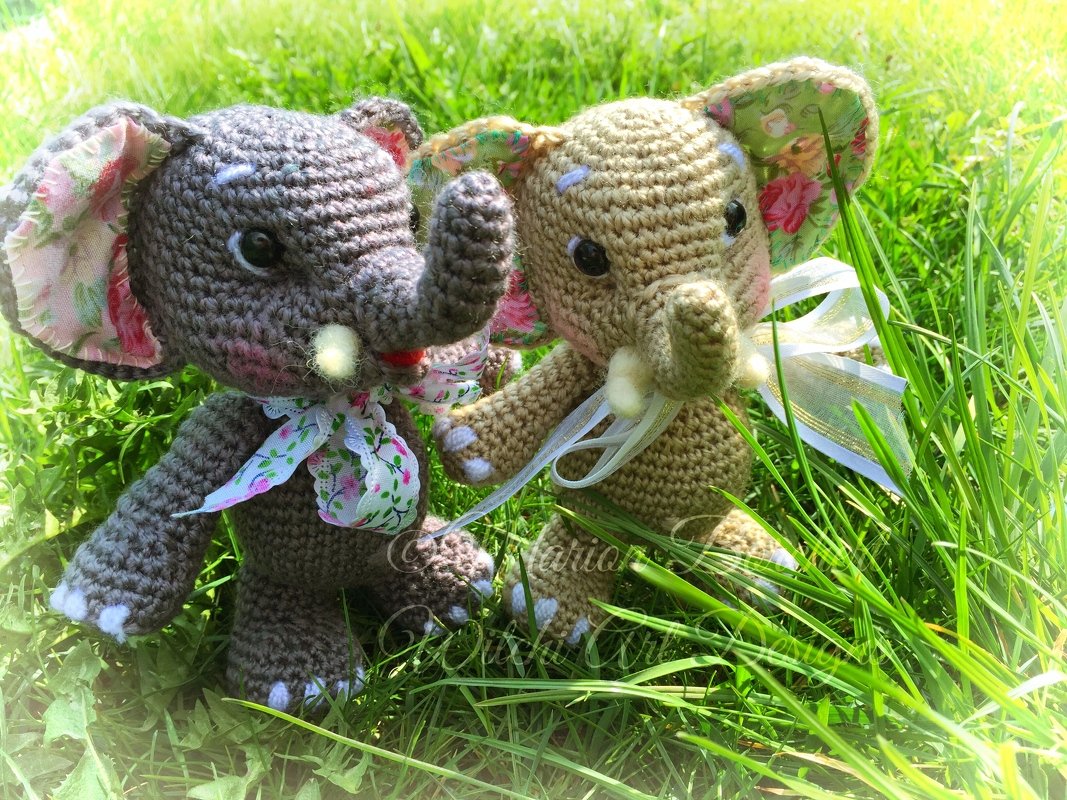 Zwei gehäkelte Elefanten-Amigurumi, grau und beige, mit floralem Ohrenfutter und Schleifen im grünen Gras.