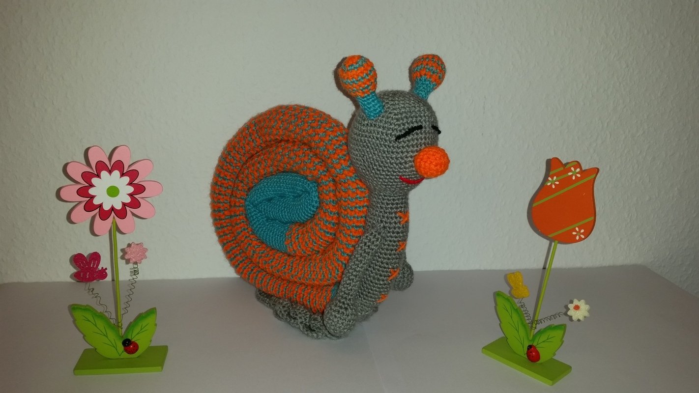 Gehäkelte Amigurumi-Schnecke mit gestreifter Spiral-Schale in Orange und Türkis neben zwei Holzblumen