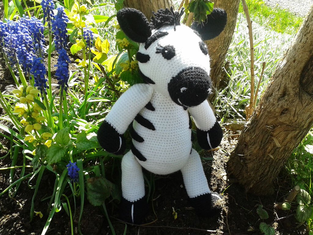 Handgehäkeltes schwarz-weißes Zebra-Amigurumi zwischen blauen Traubenhyazinthen neben einem Baumstamm