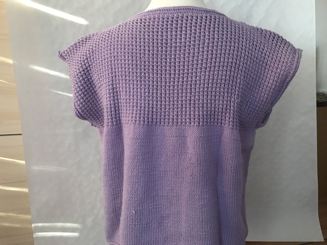 Rückansicht: ärmelloser lila Pullunder mit wabenartiger strukturierter Passe und glatt gestricktem Rumpf.