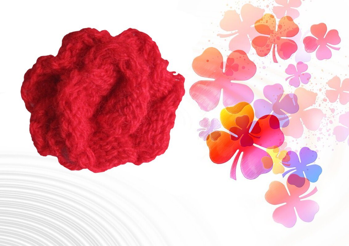 Rote gestrickte Blüte neben farbenfrohen Glückskleeblättern auf hellem Hintergrund.