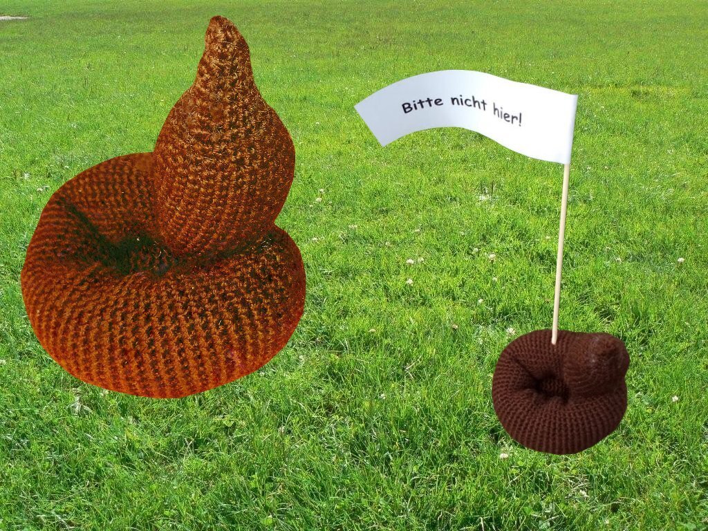 Zwei gehäkelte Hundehaufen auf Gras, einer größer braun, einer kleiner mit weißem Fähnchen ‚Bitte nicht hier!‘