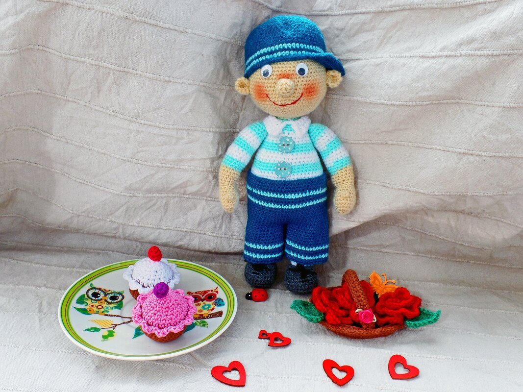 Gehäkelte Jungenpuppe in blau-weißem Outfit, umgeben von Cupcakes, Rosen, herzförmigen Dekorationen und einem Marienkäfer