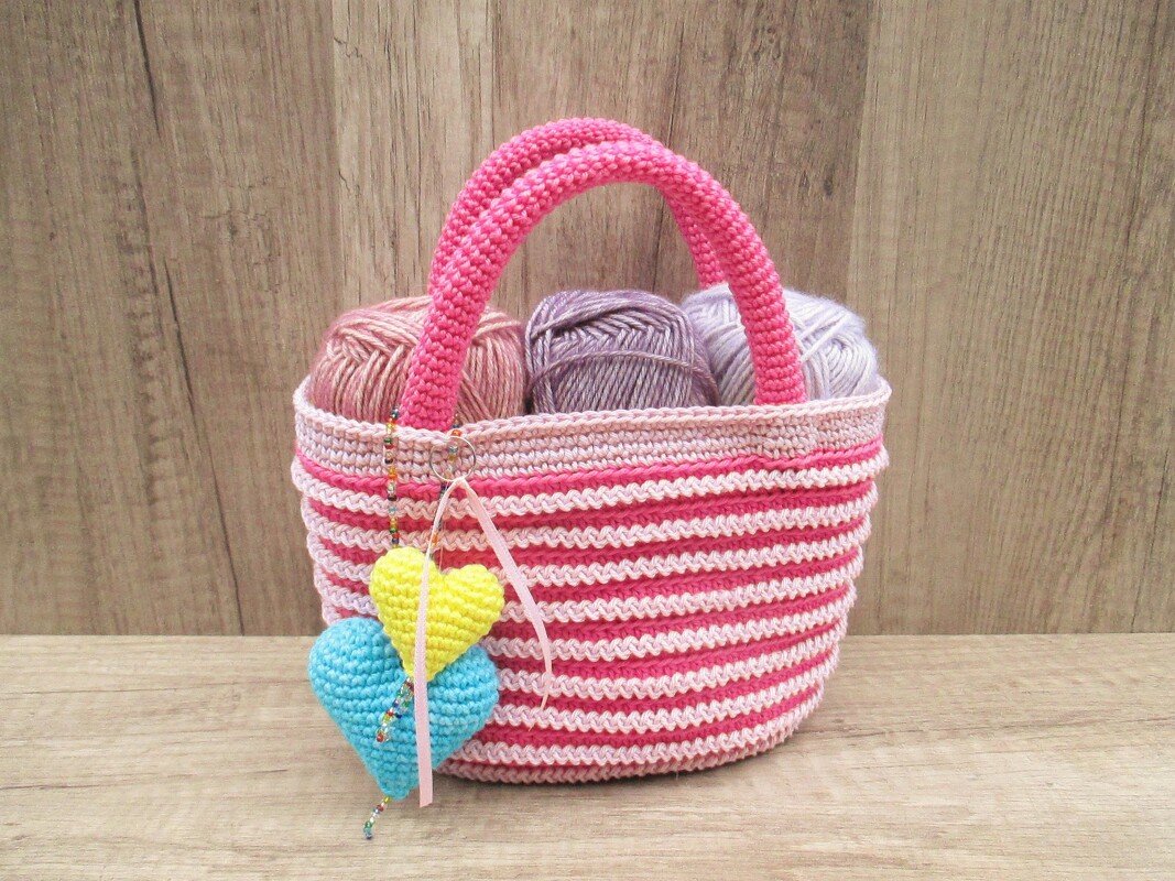 Gehäkelte Henkeltasche mit rosa Streifen, gefüllt mit Garnknäueln und zwei gehäkelten Herz-Anhängern in Gelb und Hellblau.