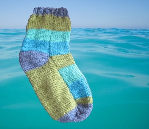 Nahaufnahme einer gestrickten Socke mit Streifen in Olivgrün, Türkis und Blau sowie lila-grauer Ferse und Spitze vor aquamarinfarbenem Wasserhintergrund.
