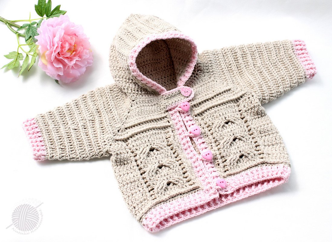 Eine gehäkelte Baby-Kapuzenjacke in Beige mit rosa Bündchen und herzförmigen Knöpfen, daneben eine rosa Blüte.