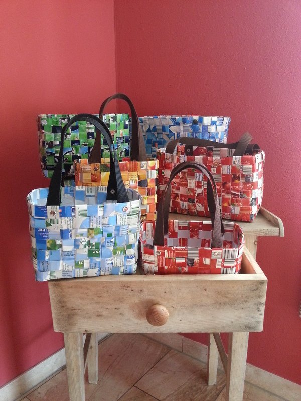 Handgeflochtene Taschen aus recycelten Getränkekartons in verschiedenen Farben auf einem Holztisch vor roter Wand