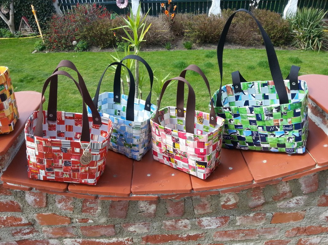 Vier bunte, geflochtene Taschen aus recycelten Getränkekartons auf einer gemauerten Mauer vor einem Garten