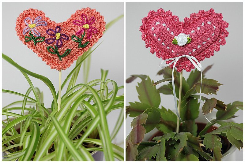 Zwei gehäkelte Herz-Pflanzenstecker: ein pfirsichfarbenes, besticktes Blumenherz und ein rosafarbenes Herz mit weißem Band, in Zimmerpflanzen gesteckt.