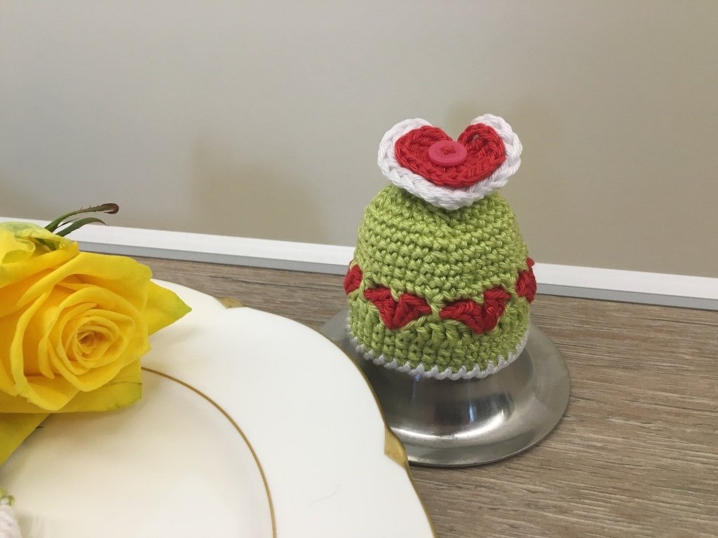 Hellgrüner gehäkelter Eierwärmer mit roten Herzapplikationen und Knopf auf Metallständer neben gelber Rose und Teller