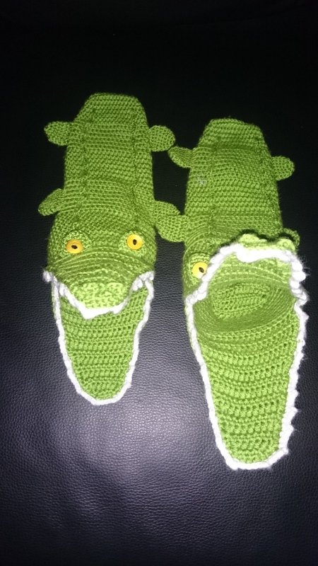 Hellgrüne gehäkelte Krokodil-Pantoffeln mit gelben Knopfaugen und weißer Umrandung auf schwarzem Hintergrund.