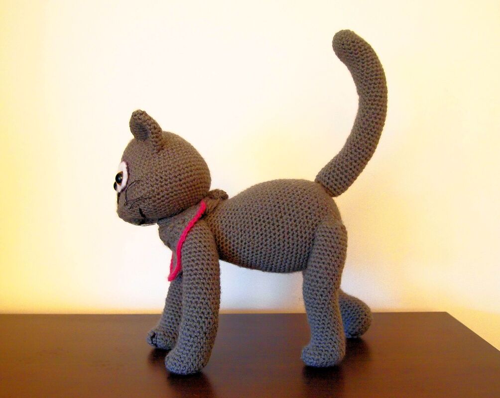 Gehäkelte graue Amigurumi-Katze mit rosa Halstuch, seitliche Ansicht, stehende Pose