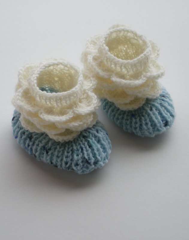 Zwei gestrickte Babyschuhe in Hellblau und Creme mit Rüschen auf weißem Hintergrund.