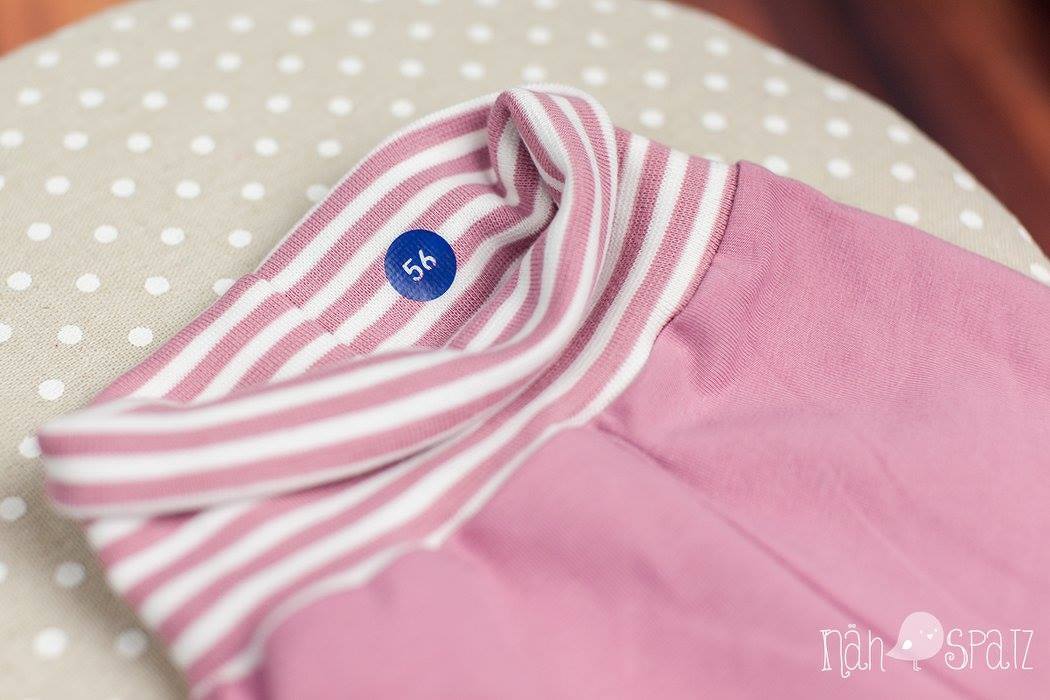 Nahaufnahme eines gestreiften Bündchens an einem rosa Babykleidungsstück mit einem blauen Aufkleber '56'.