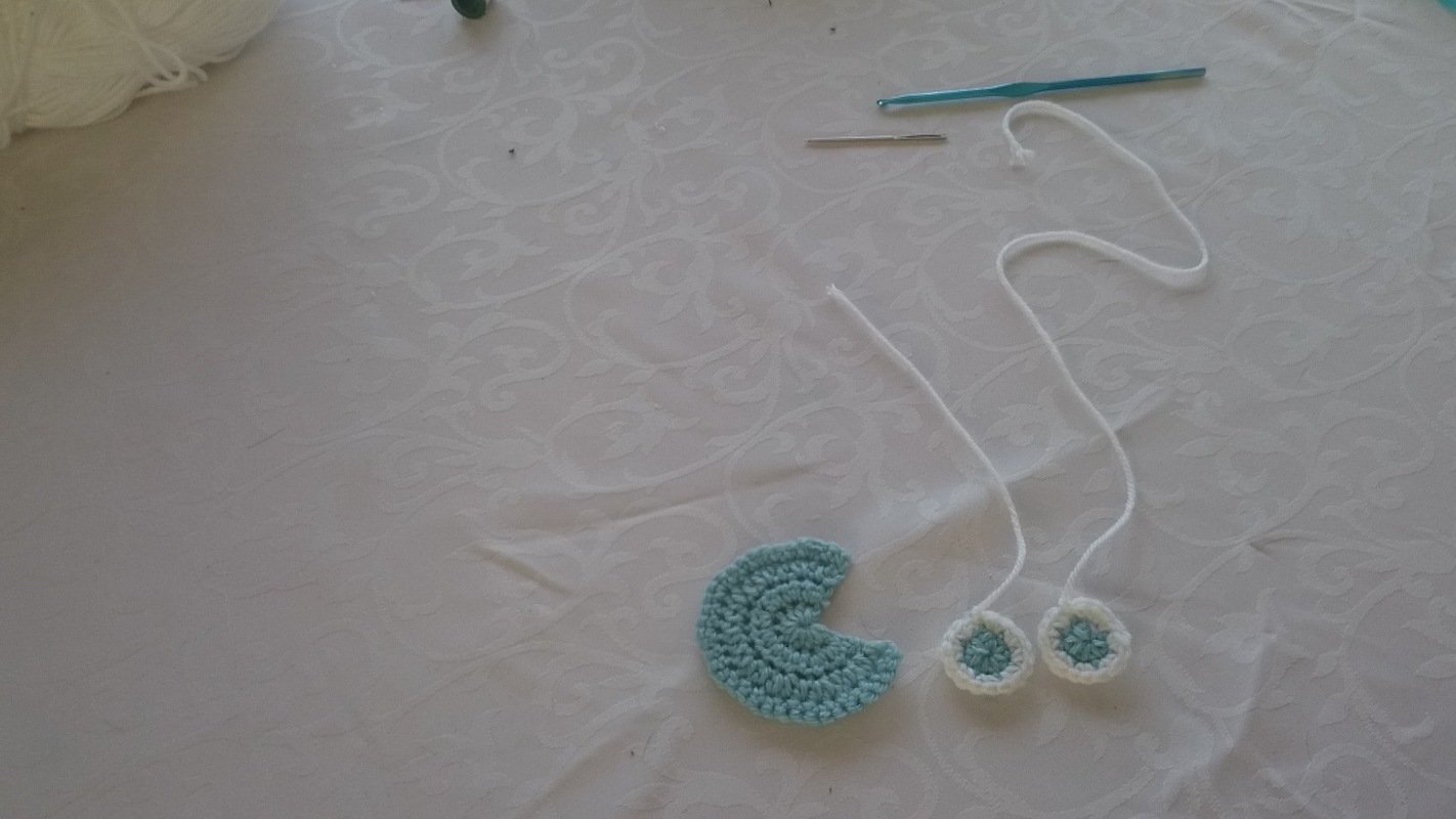 Hellblaues gehäkeltes Halbmondstück, zwei kleine runde Applikationen mit weißen Schnüren, Knäuel weiße Wolle, blaue Häkelnadel und Stopfnadel auf weißem Tischtuch