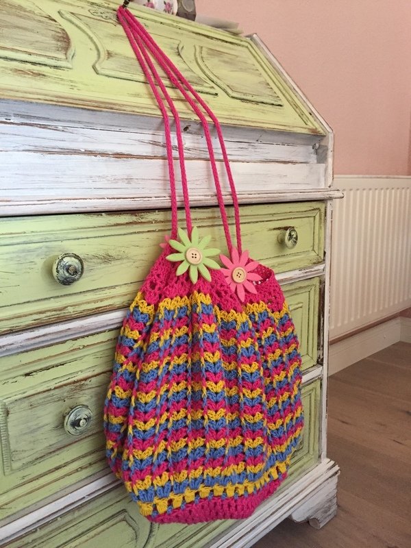 Gehäkelte Umhängetasche in Pink, Gelb und Blau mit Blumenknöpfen, hängt an einer grünen Kommode.