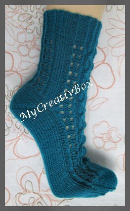 Petrolfarbene gestrickte Socke mit Zopf- und Lochmuster auf hellem Untergrund.