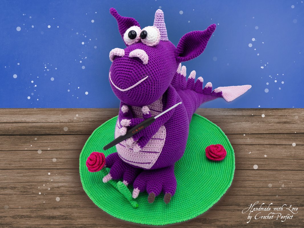 Lila Amigurumi-Drache mit großen Augen hält eine Häkelnadel und sitzt auf einer runden grünen Matte neben roten Rosen.