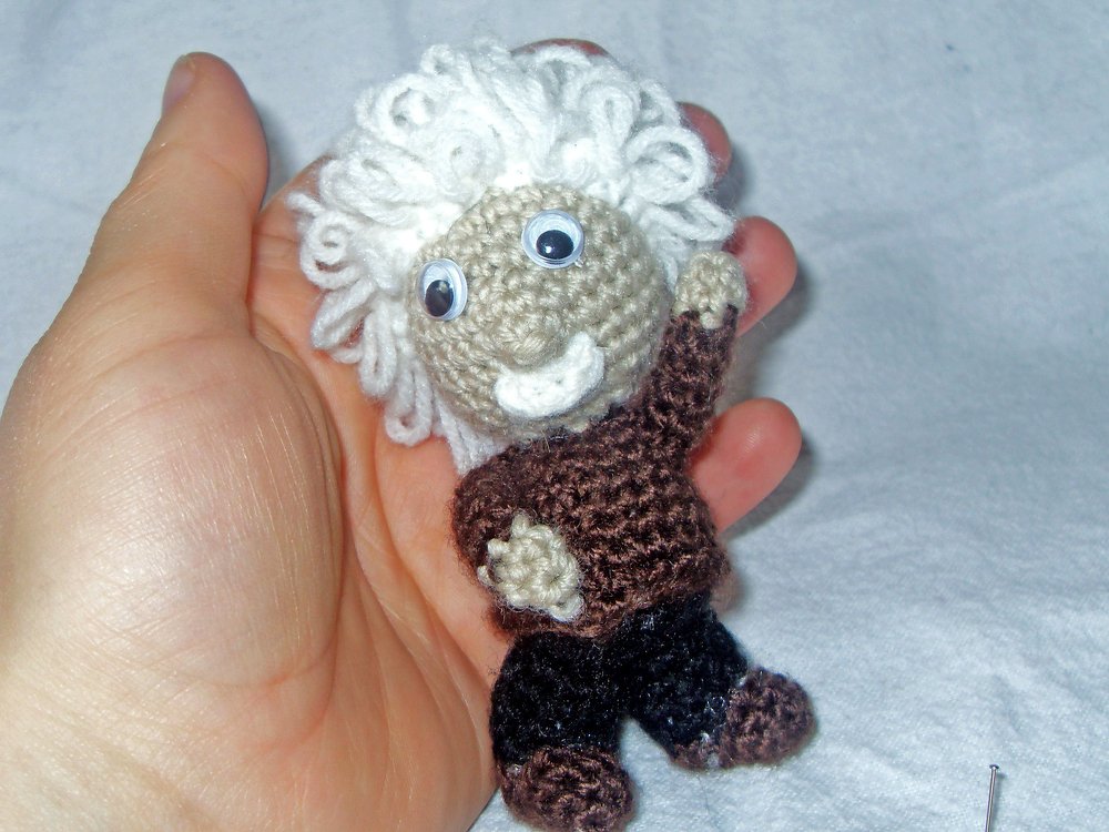 Hand hält kleines gehäkeltes Amigurumi mit weißen Locken und braunem Oberteil.